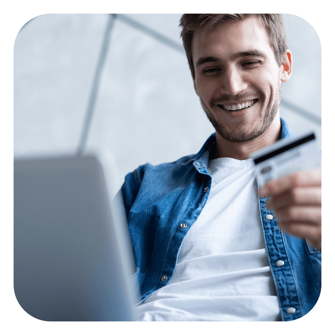 Uma pessoa sorridente, vestindo uma camisa jeans, segura um cartão de crédito enquanto olha para a tela de um laptop.