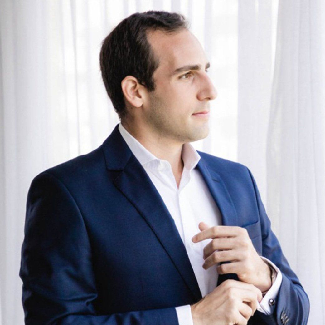 Leonardo Rembischewiski, CEO da RK2 Seguros, de terno azul e camisa branca ajusta o punho da camisa, em pé em frente a cortinas brancas transparentes.