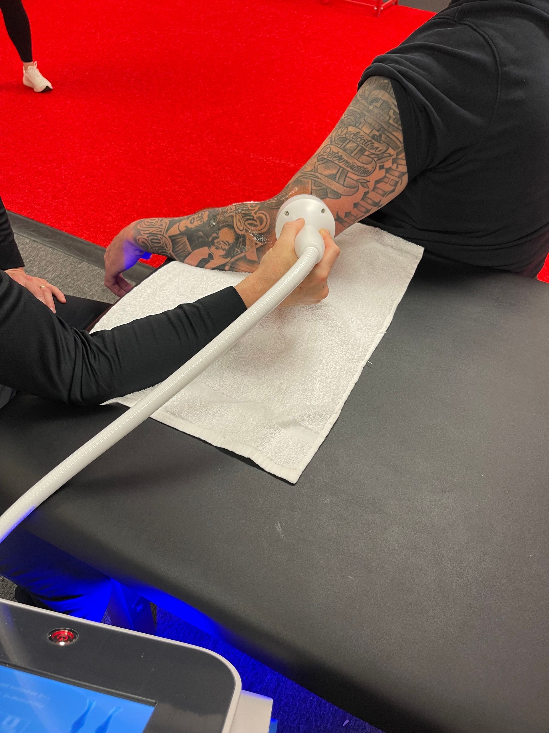 Person receiving treatment on their arm, using a handheld device on a white towel, on a black table, red floor.