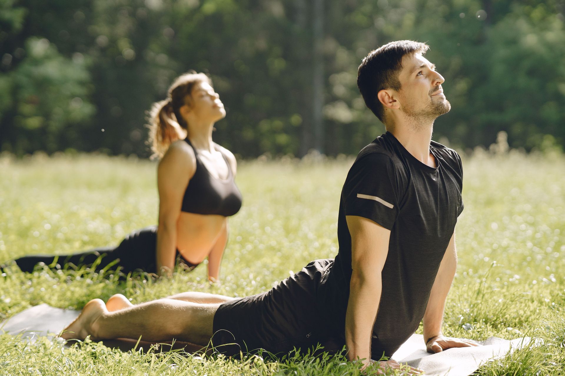 Two people doing yoga on mats in a sunny grassy park, one in a cobra pose.