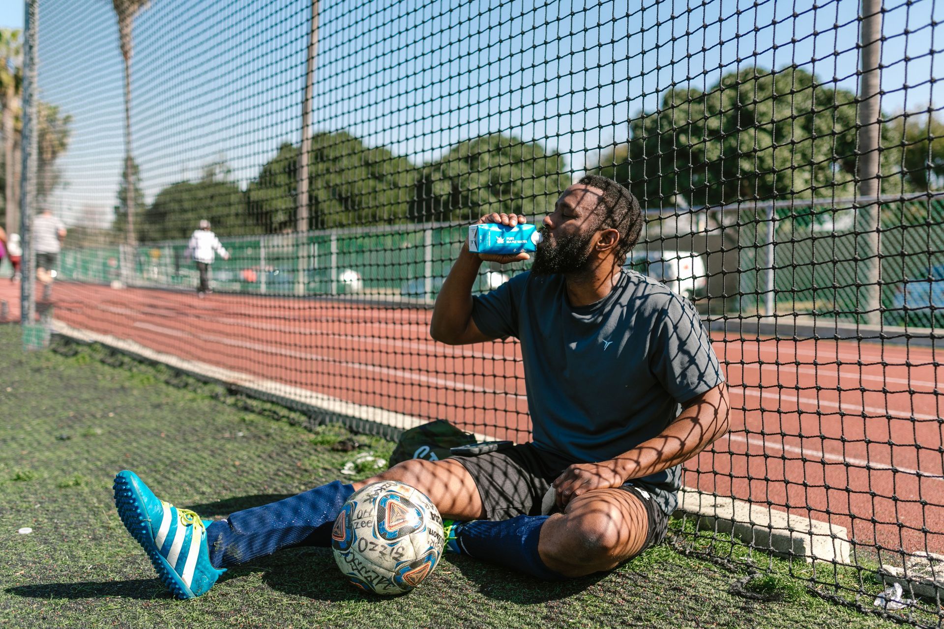 Soccer player sitting on a field, drinking water beside a soccer ball near a goal net.