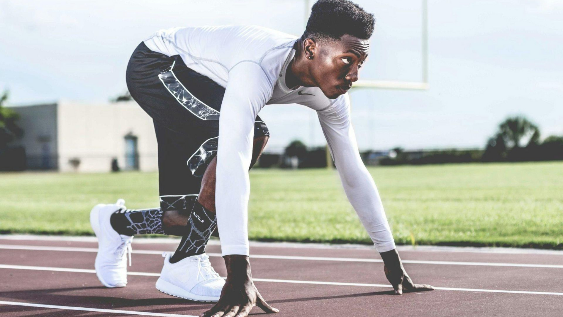 A sprinter in a white long-sleeve shirt and black athletic shorts crouches in a starting position on a running track.