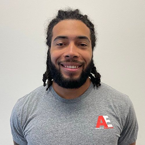 A man with dreadlocks and a beard is wearing a grey t-shirt and smiling.
