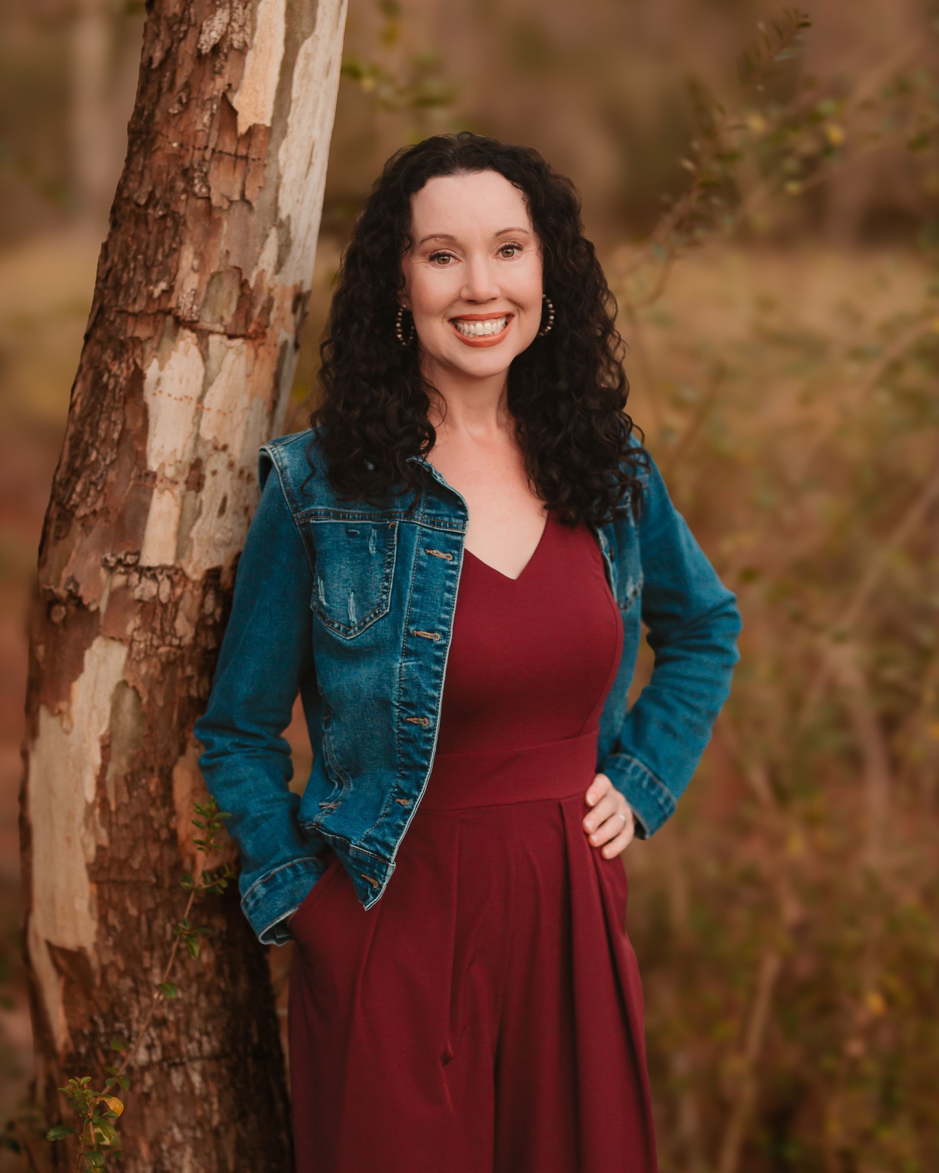 A woman in a red dress and denim jacket is standing next to a tree.