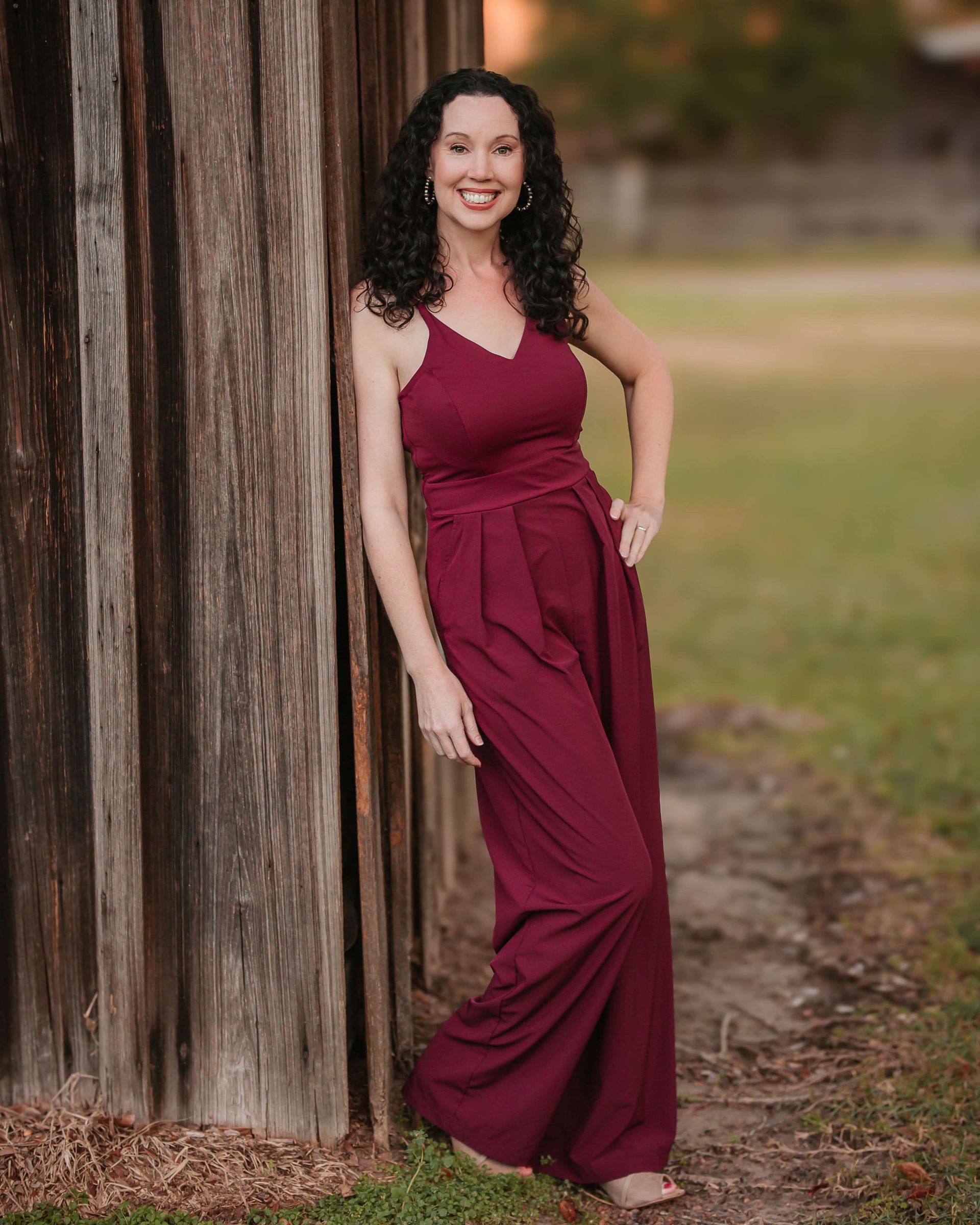 A woman in a maroon jumpsuit is leaning against a wooden fence.