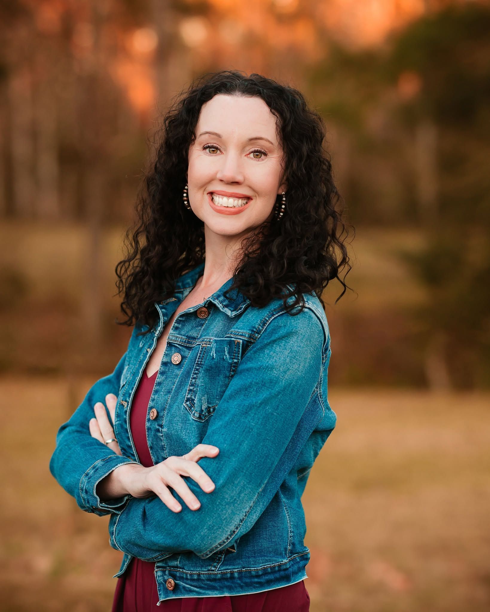 A woman in a denim jacket is standing in a field with her arms crossed and smiling.