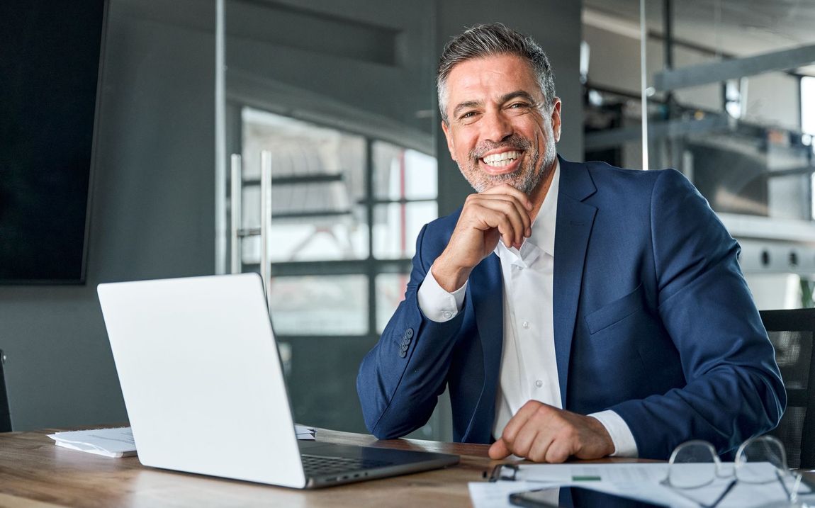 A man in a suit is sitting at a desk in front of a laptop computer.