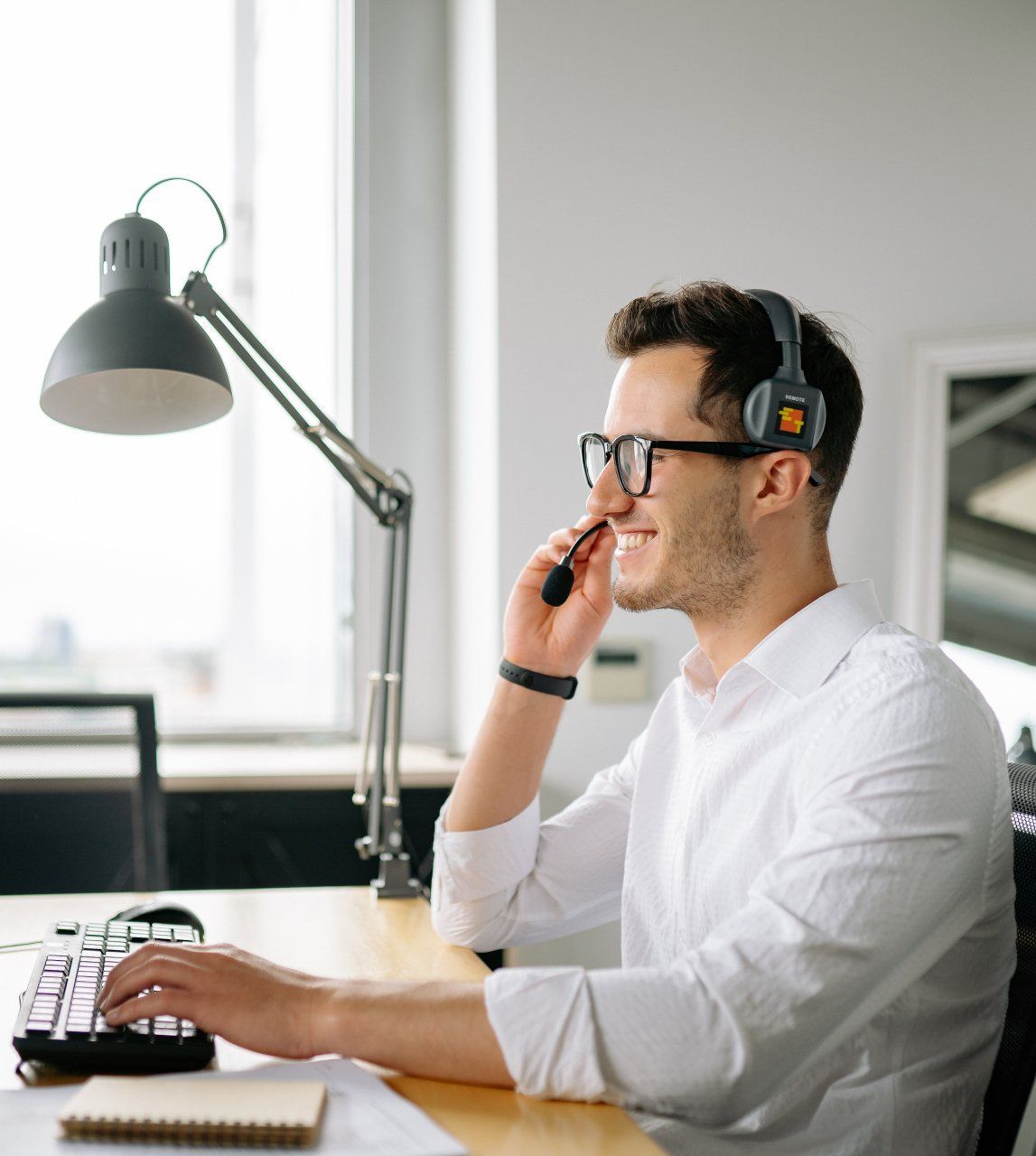 A man wearing headphones is talking on a cell phone while typing on a keyboard