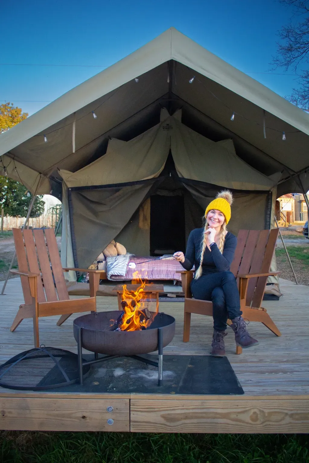 Woman in yellow hat sits by fire pit in front of a glamping tent, smiling.