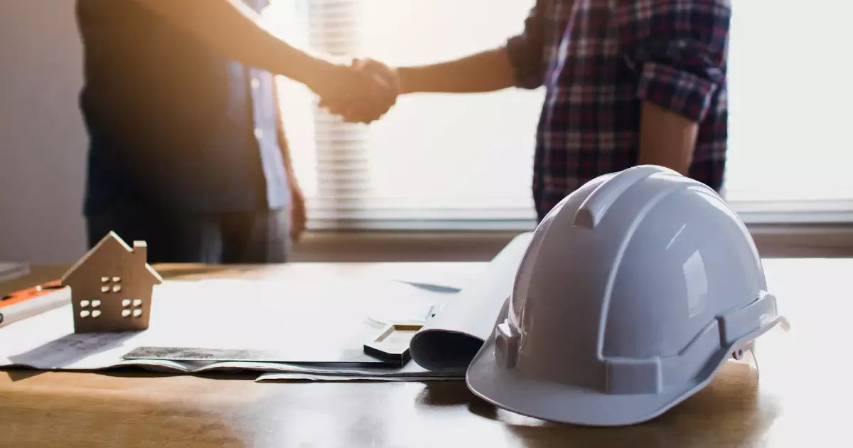 Builders shaking hands over blueprints with safety helmet and house model on table during custom home building agreement