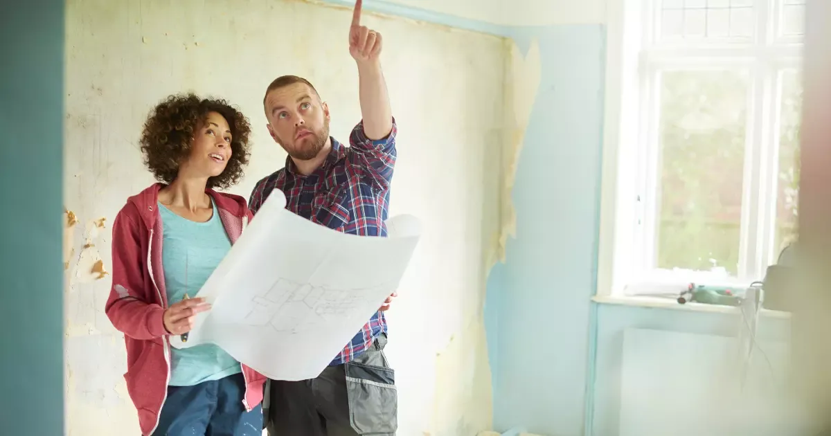 Couple reviewing blueprints with contractor inside unfinished room during home renovation project