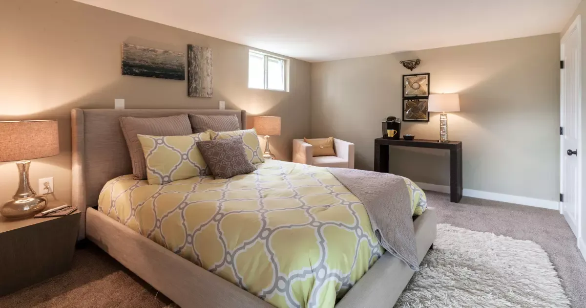 Cozy basement bedroom with plush carpet, warm lighting, queen bed, and small window for natural light