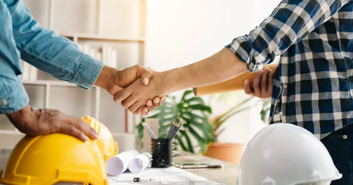 Contractor and client shaking hands over blueprints and hard hats during custom home building project planning