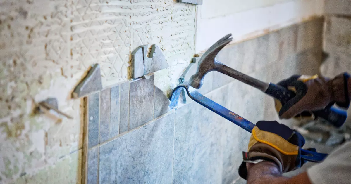 Worker removing old wall tiles with hammer and pry bar during interior home construction renovation