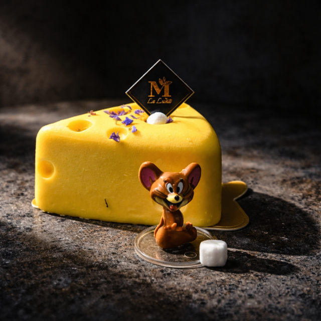 Yellow cheese wedge dessert with purple flowers and a small mouse figurine on a dark surface