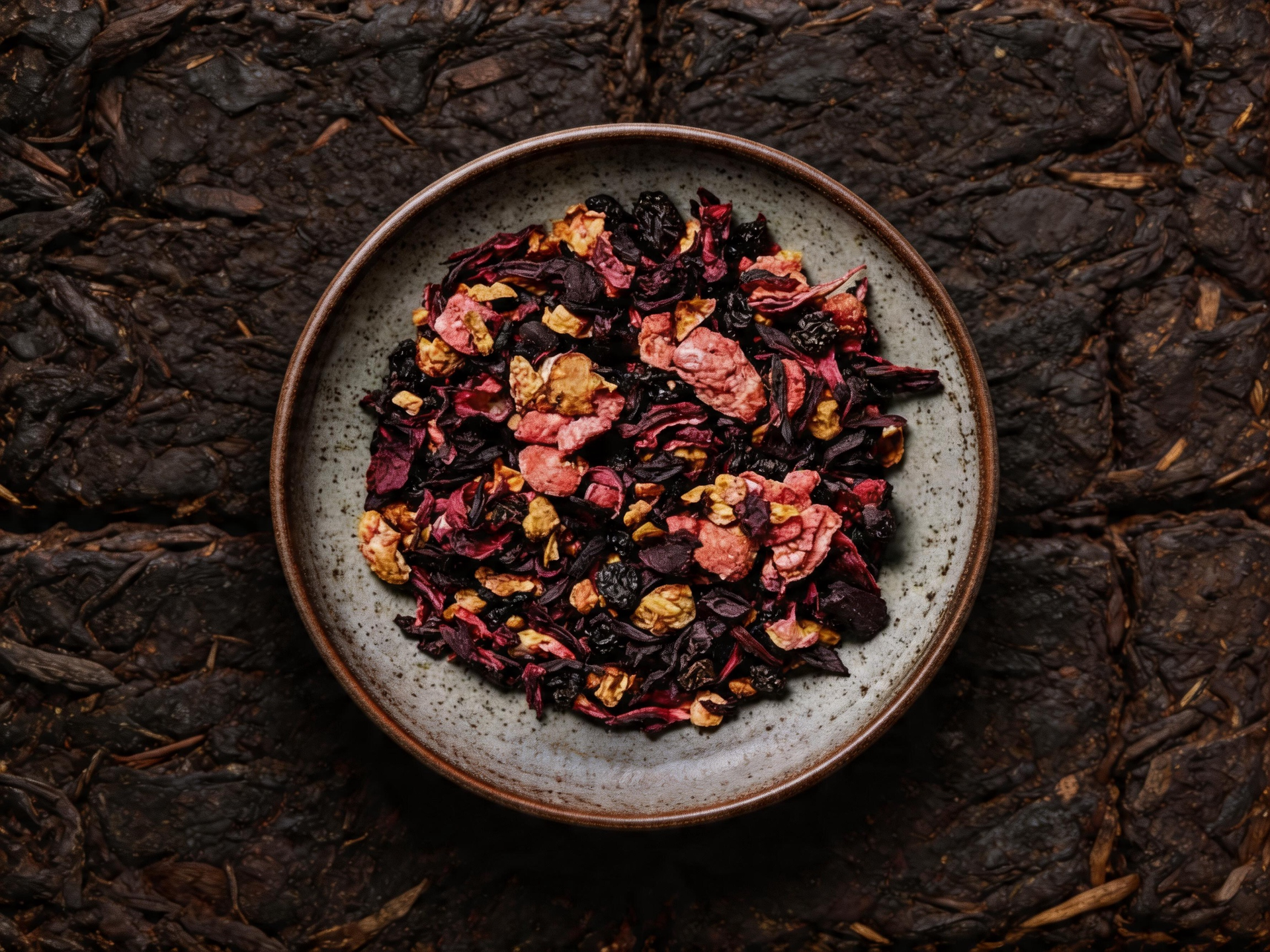 Top-down view of a bowl of dried flower petals on a dark textured surface