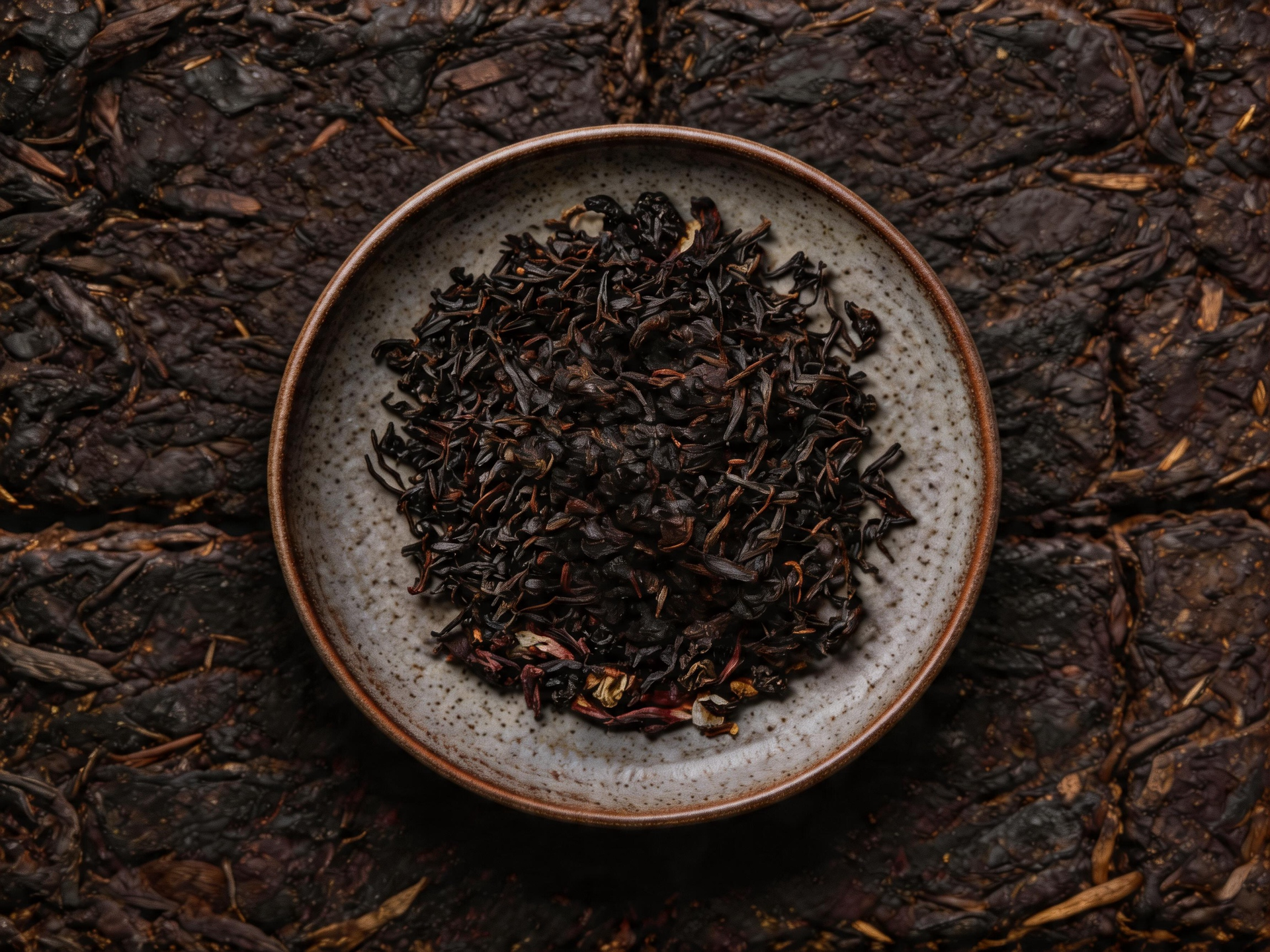 A bowl of loose dark tea leaves on a textured dark surface.