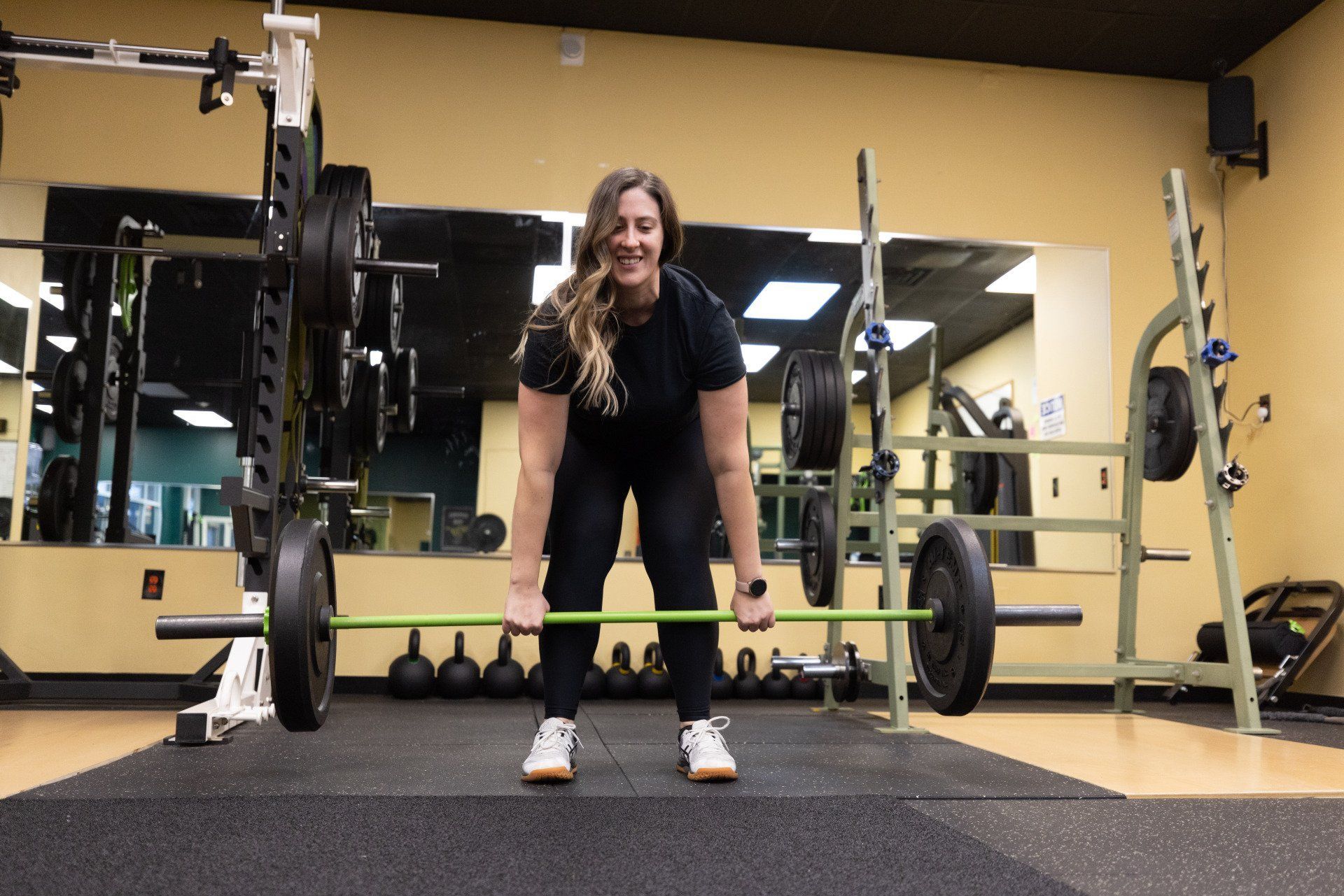 A person standing in a gym, smiling while holding a barbell with black weights in a deadlift starting position.