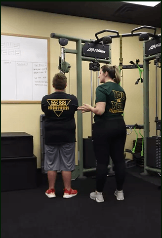 A trainer instructs a client on how to use a cable machine in a gym, with a whiteboard on the wall nearby.