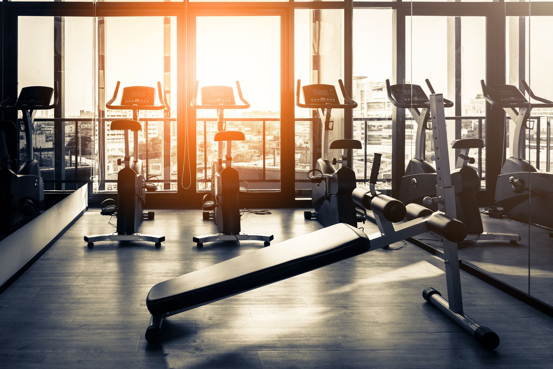 An empty gym featuring rows of stationary exercise bikes and a workout bench, illuminated by bright, warm sunlight.