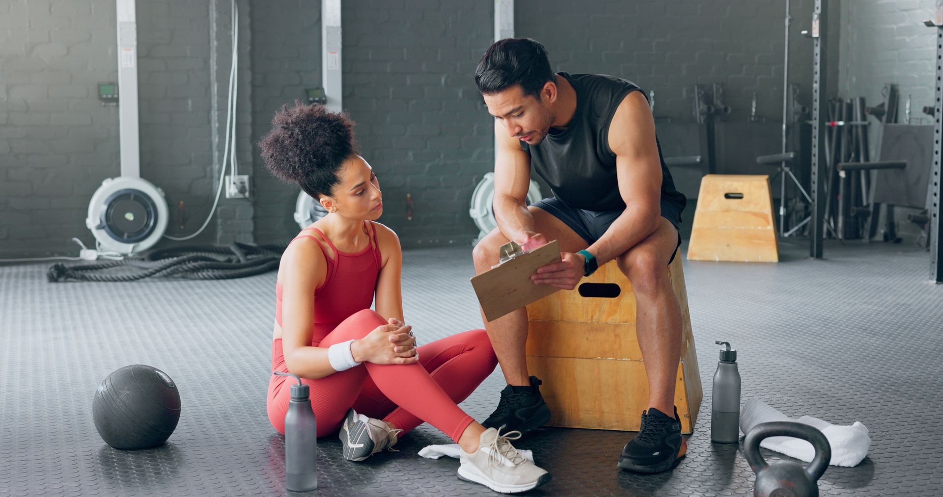 A personal trainer reviews a clipboard with a client sitting on the floor in a gym.