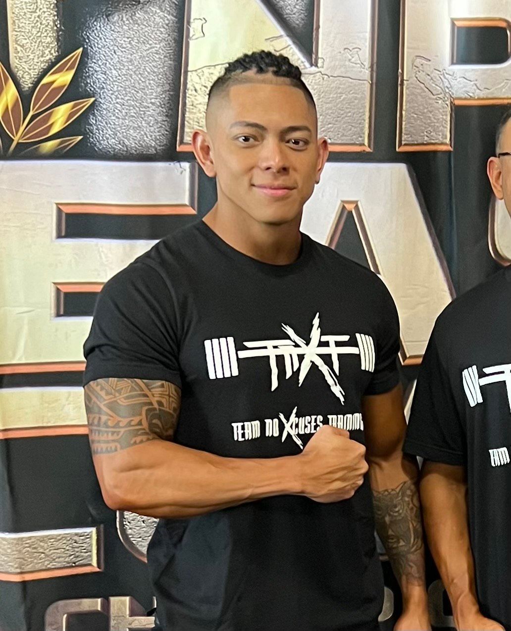 A smiling person in a black graphic t-shirt poses with a closed fist against a backdrop with metallic gold text.