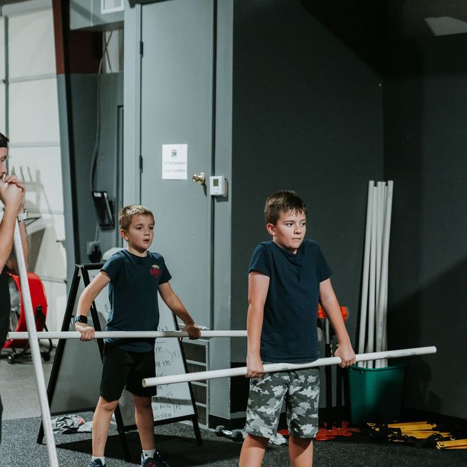 Two children holding barbells in a gym, standing side by side near a mirror.