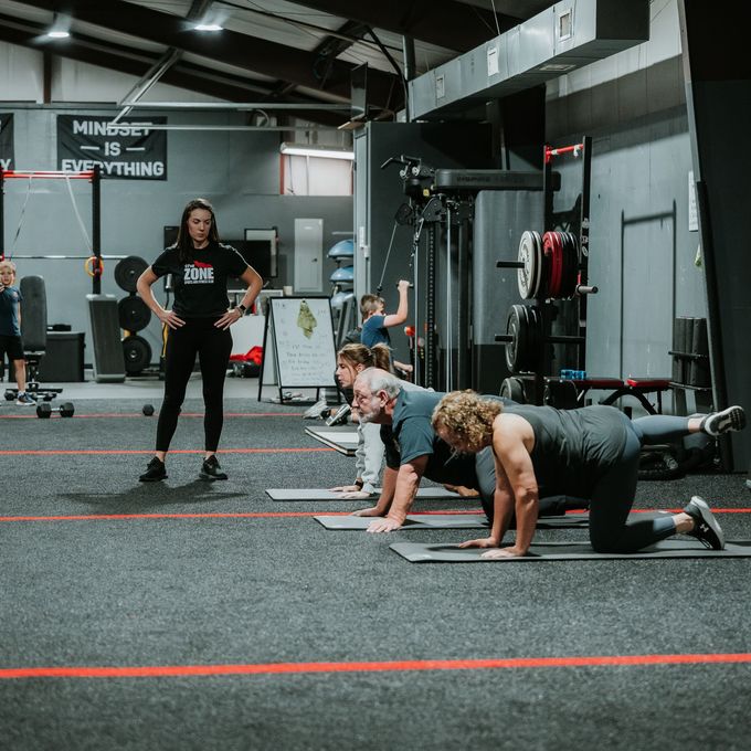 People exercising in a gym, with two doing plank push-ups and a coach standing nearby.