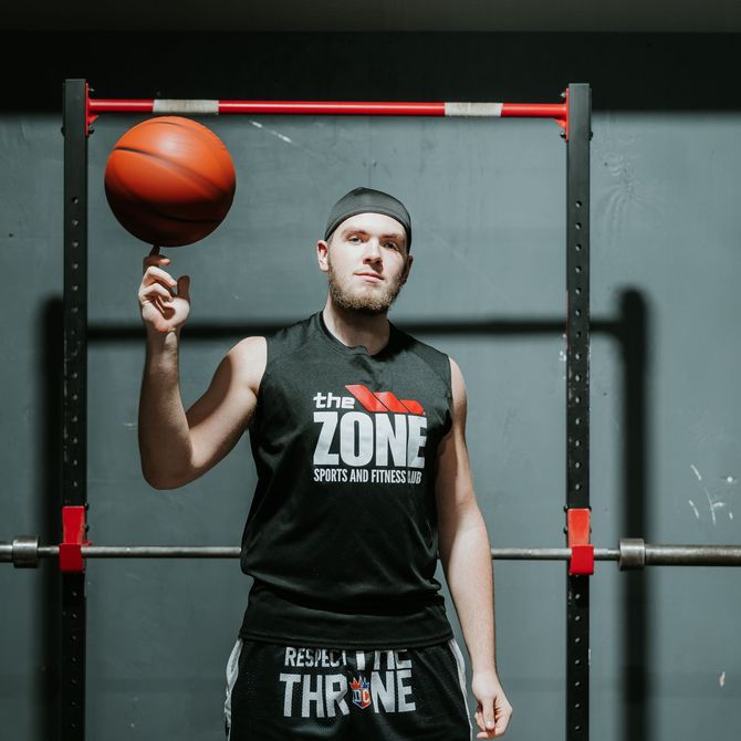 Man spinning a basketball on one finger in a gym, standing before a bar rack, wearing a black sleeveless shirt.