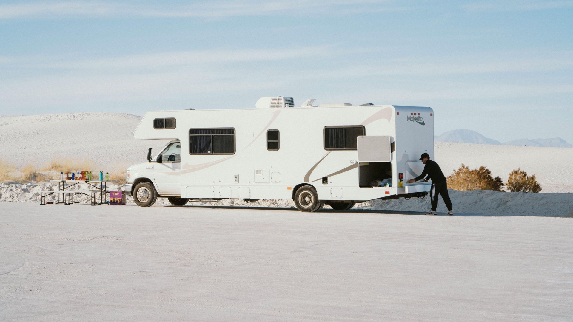 White RV parked on a snowy plain, with a person pushing at the rear near distant mountains.