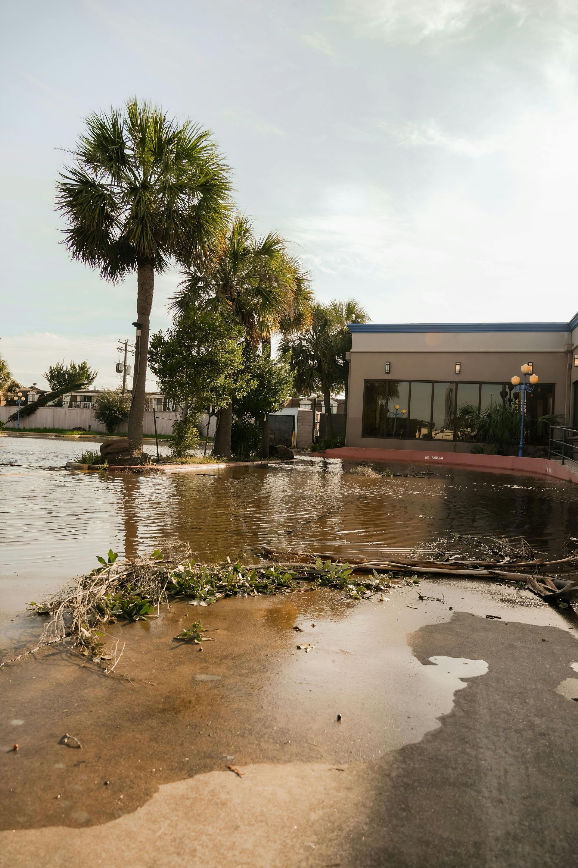 Flooded parking lot with standing water reflecting a building and trees under a cloudy sky