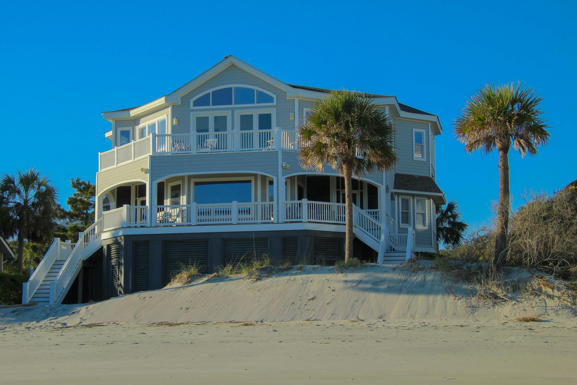 Beachfront house with white trim and palm trees on sandy dunes under a clear blue sky