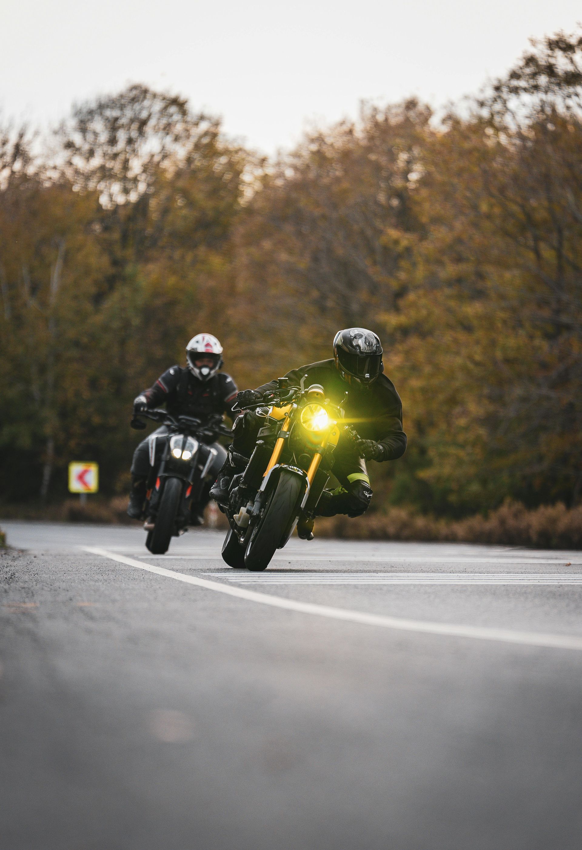 Two motorcyclists lean into a curve on a wet road, one with a bright yellow headlight.