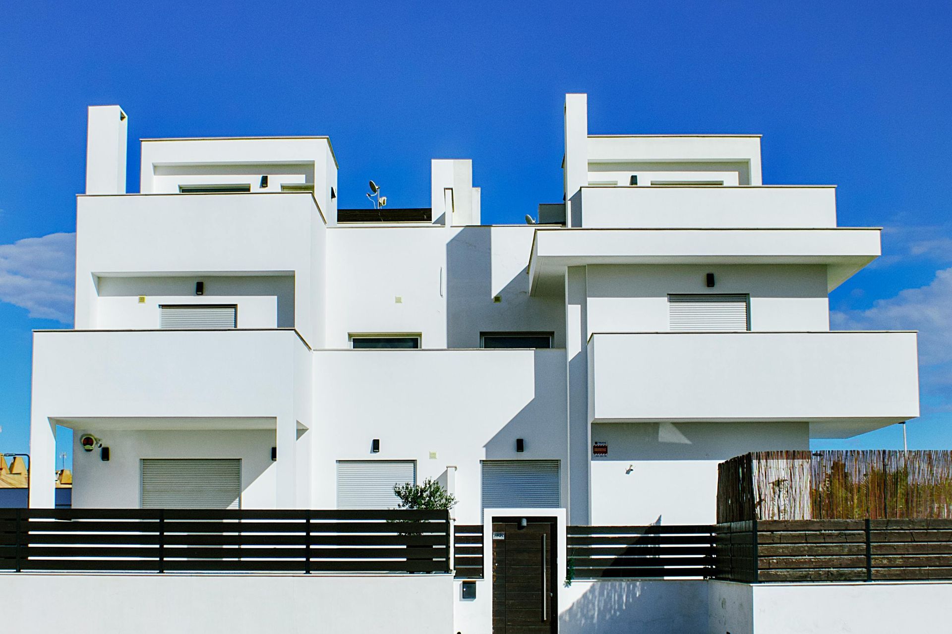 Modern white multi-story house with balconies and a black fence under a clear blue sky
