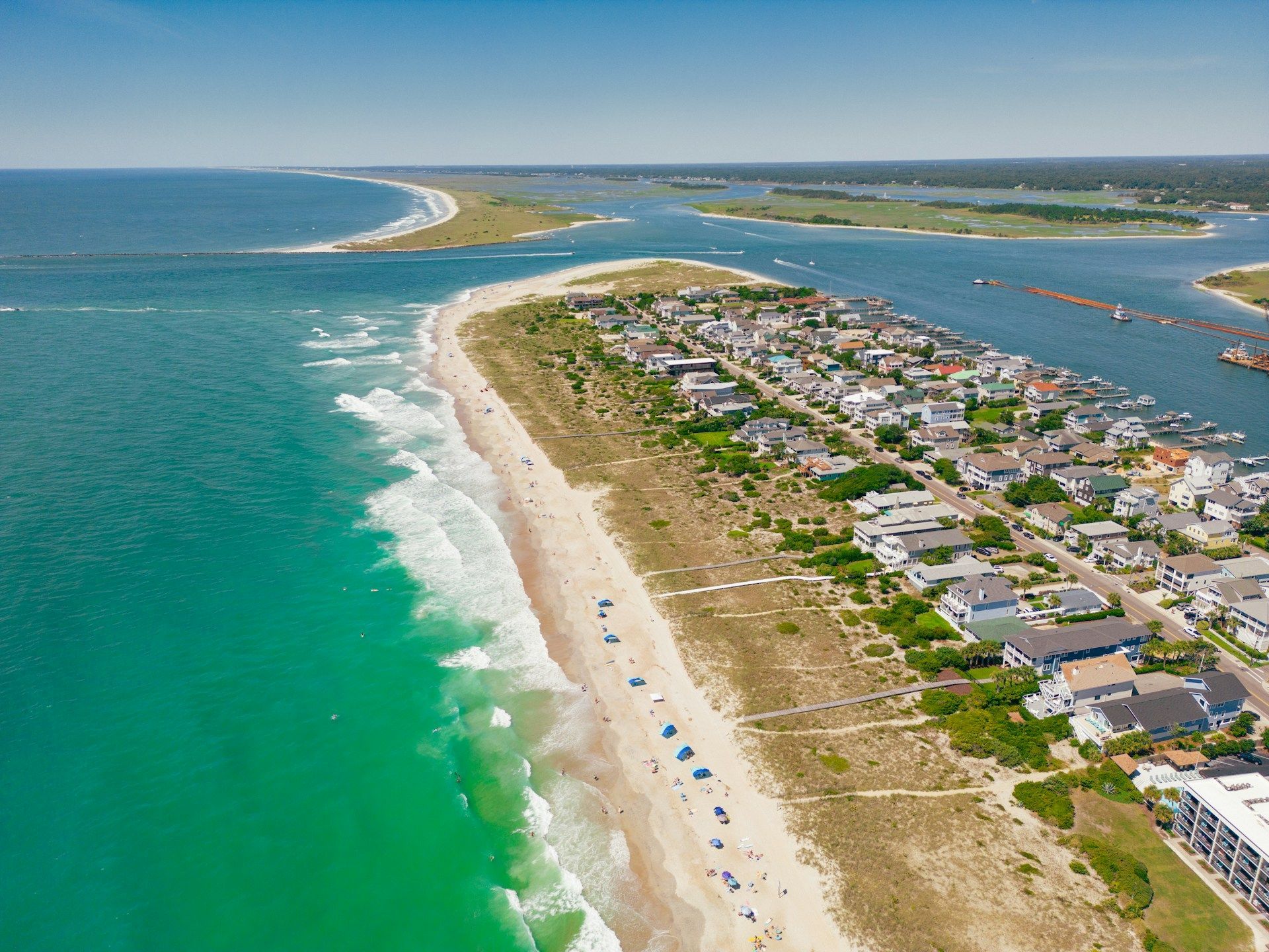 Aerial view of a turquoise beach beside a coastal town and waterway, with dunes and rows of houses.