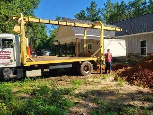 Yellow truck with crane next to a house under construction; a worker stands nearby.