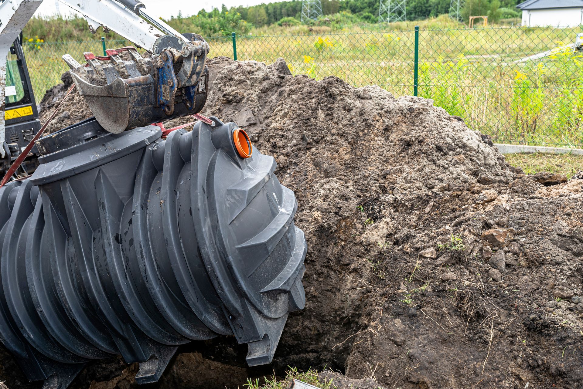 Excavator placing a dark septic tank in a hole next to a pile of dirt and a green fence.