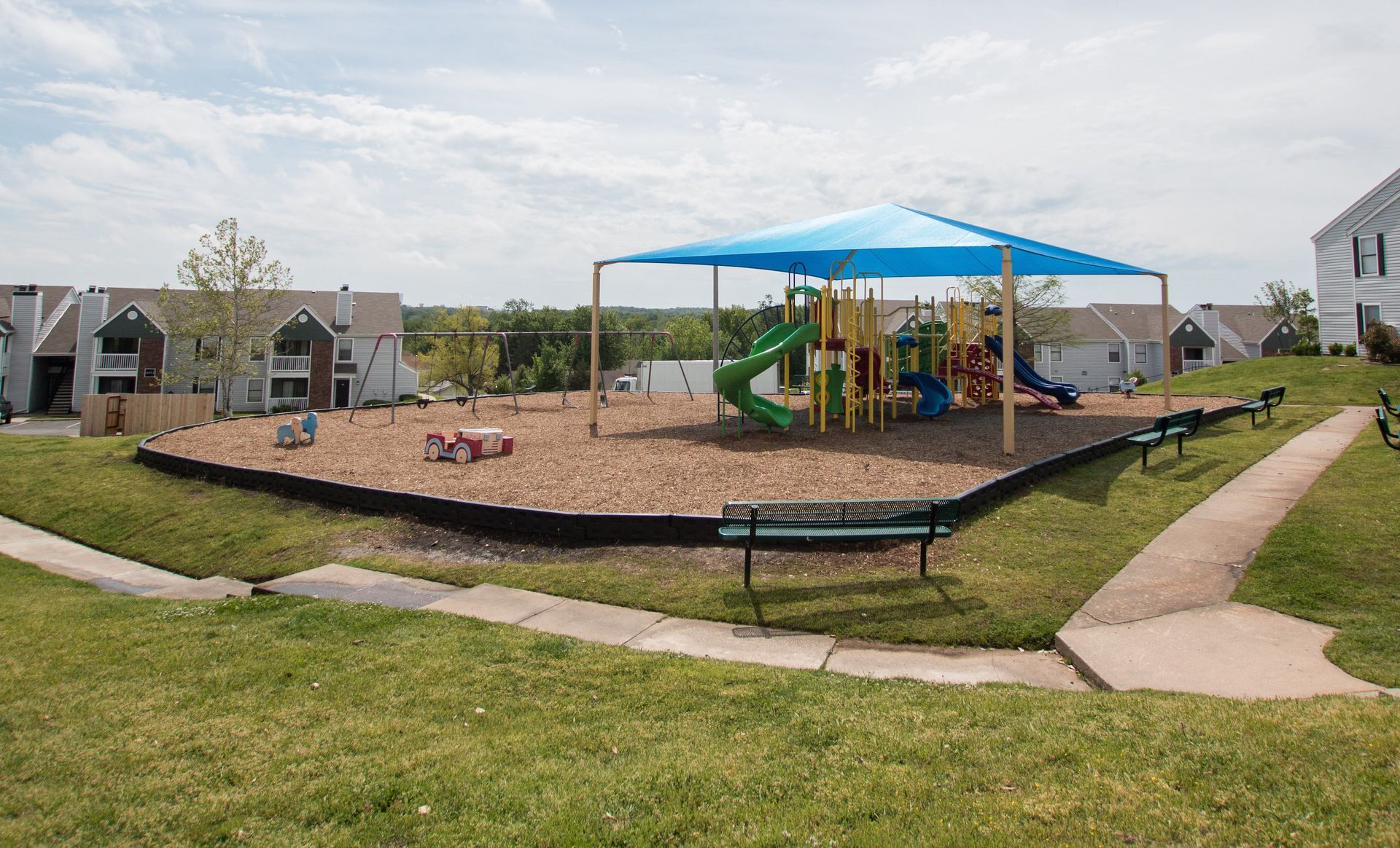 A playground with a blue umbrella and a bench in front of a house.