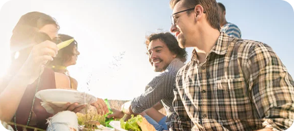 Group of people smiling, gathering outdoors. Sunlight, food, plaid shirts, clear sky.