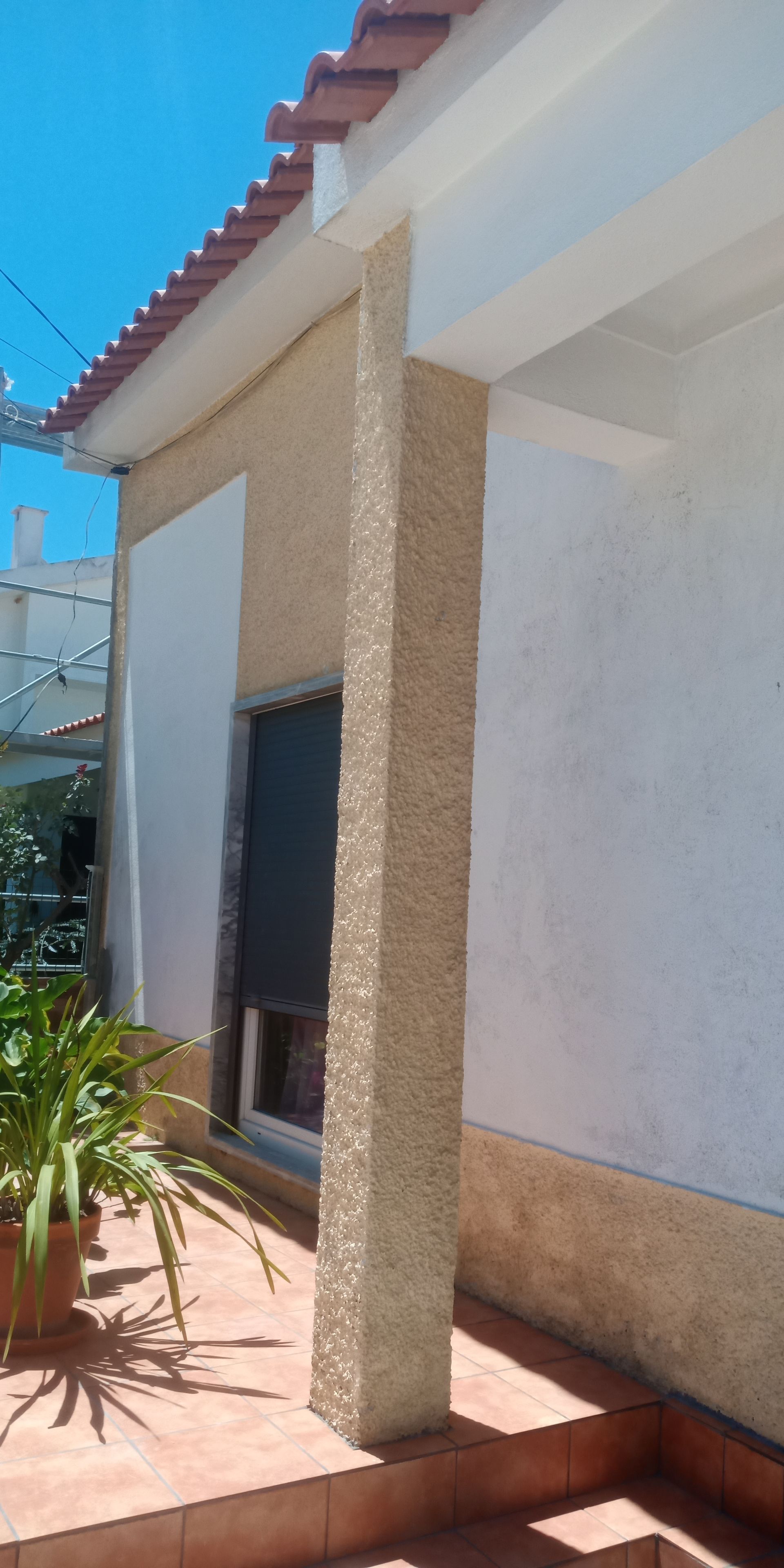Exterior de um edifício com uma coluna texturizada, uma janela e um pátio de ladrilhos de terracota sob um céu azul.