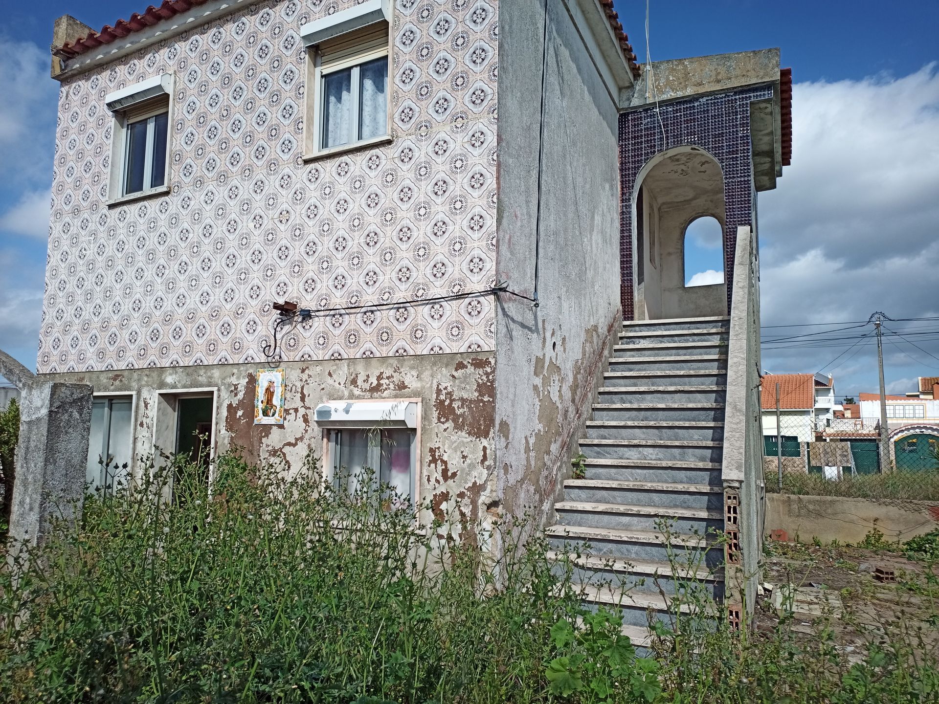 Prédio de dois andares com fachada padronizada, escadas que levam a uma entrada em arco e vegetação exuberante em primeiro plano.