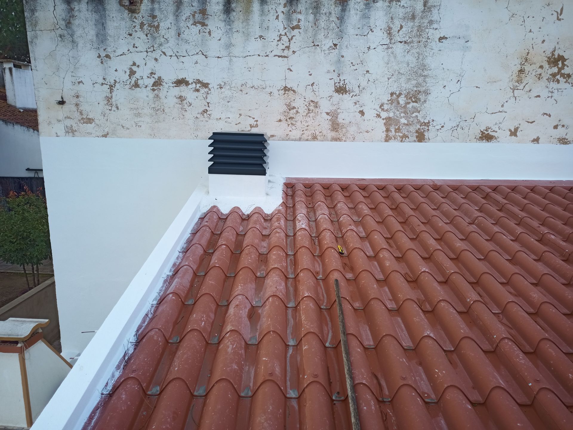 Telhado de telha vermelha com chaminé preta encostado em uma parede branca descascada.