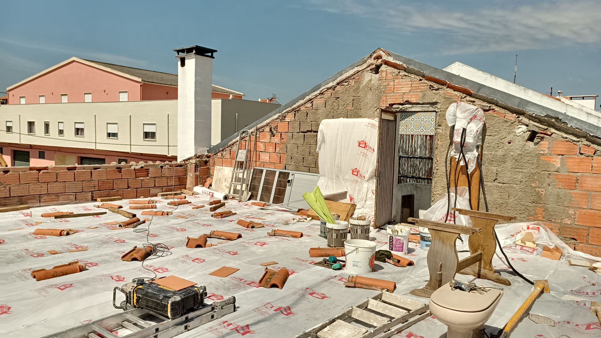 Telhado em construção: tijolos aparentes, lonas, telhas espalhadas, ferramentas e uma vista parcial dos edifícios.