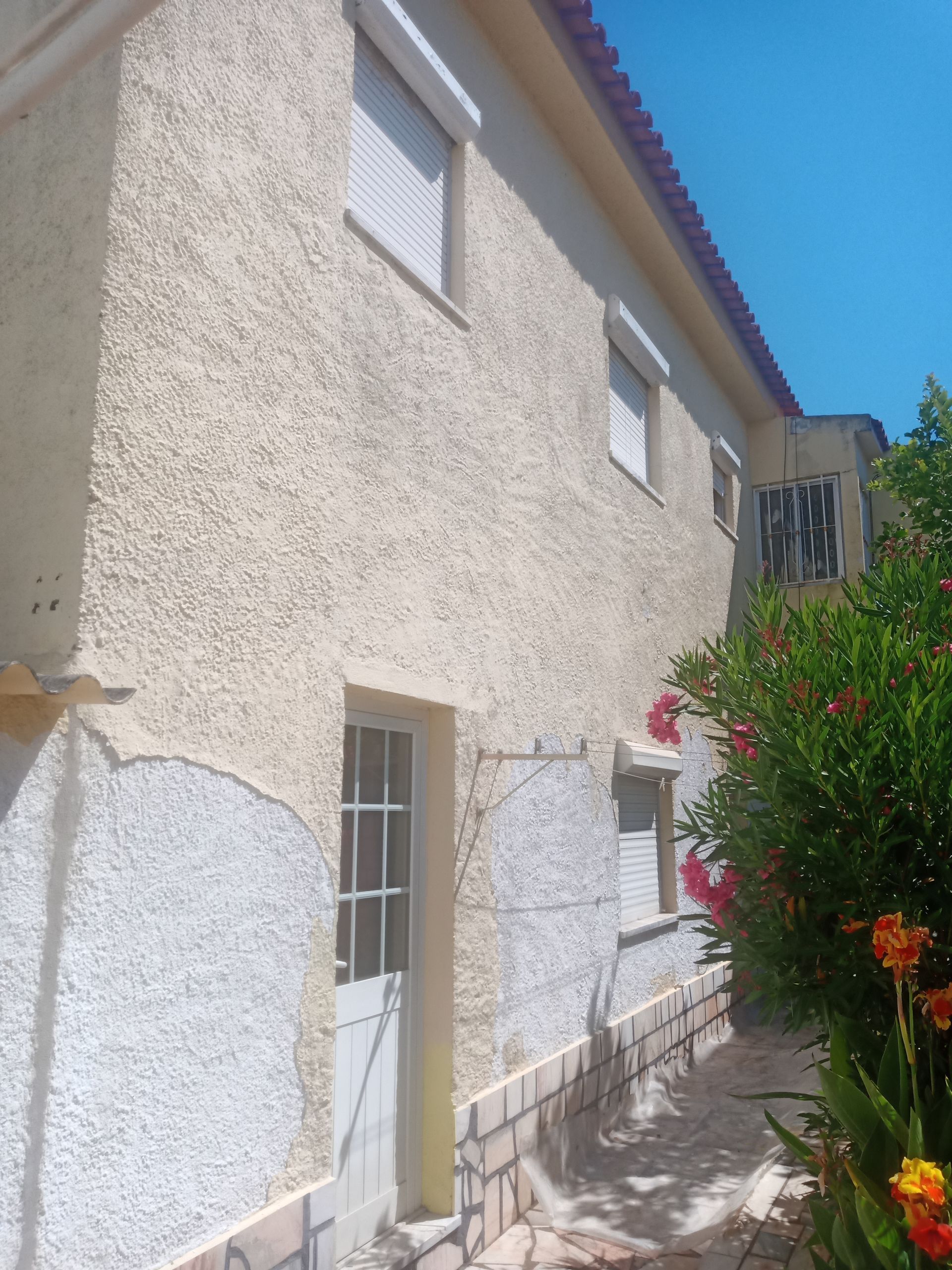 Prédio de dois andares com paredes texturizadas em tom creme, janelas e venezianas brancas, porta e telhado de telhas vermelhas.
