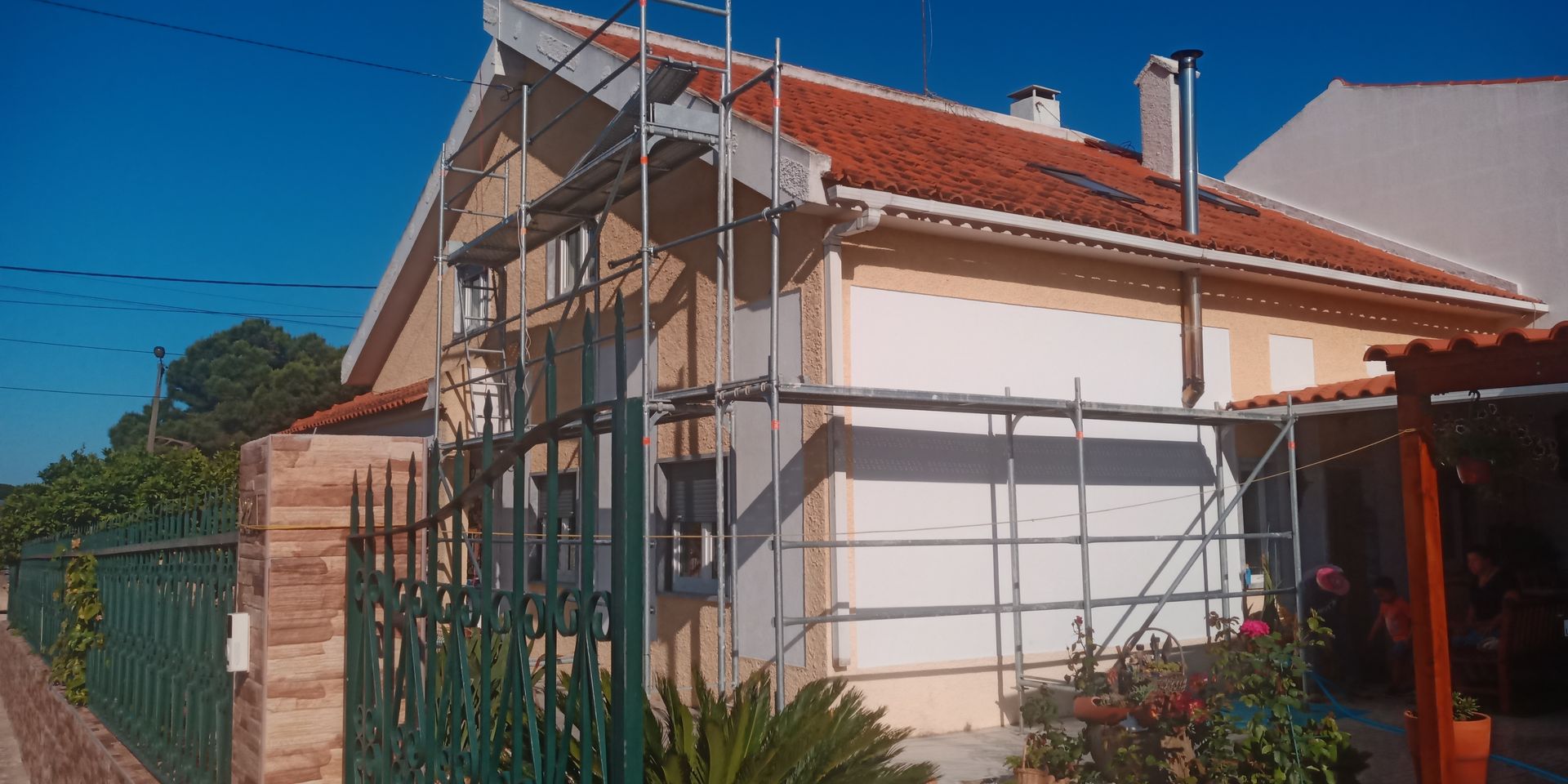 Casa com telhado de telha vermelha, andaimes e cerca verde contra um céu azul.