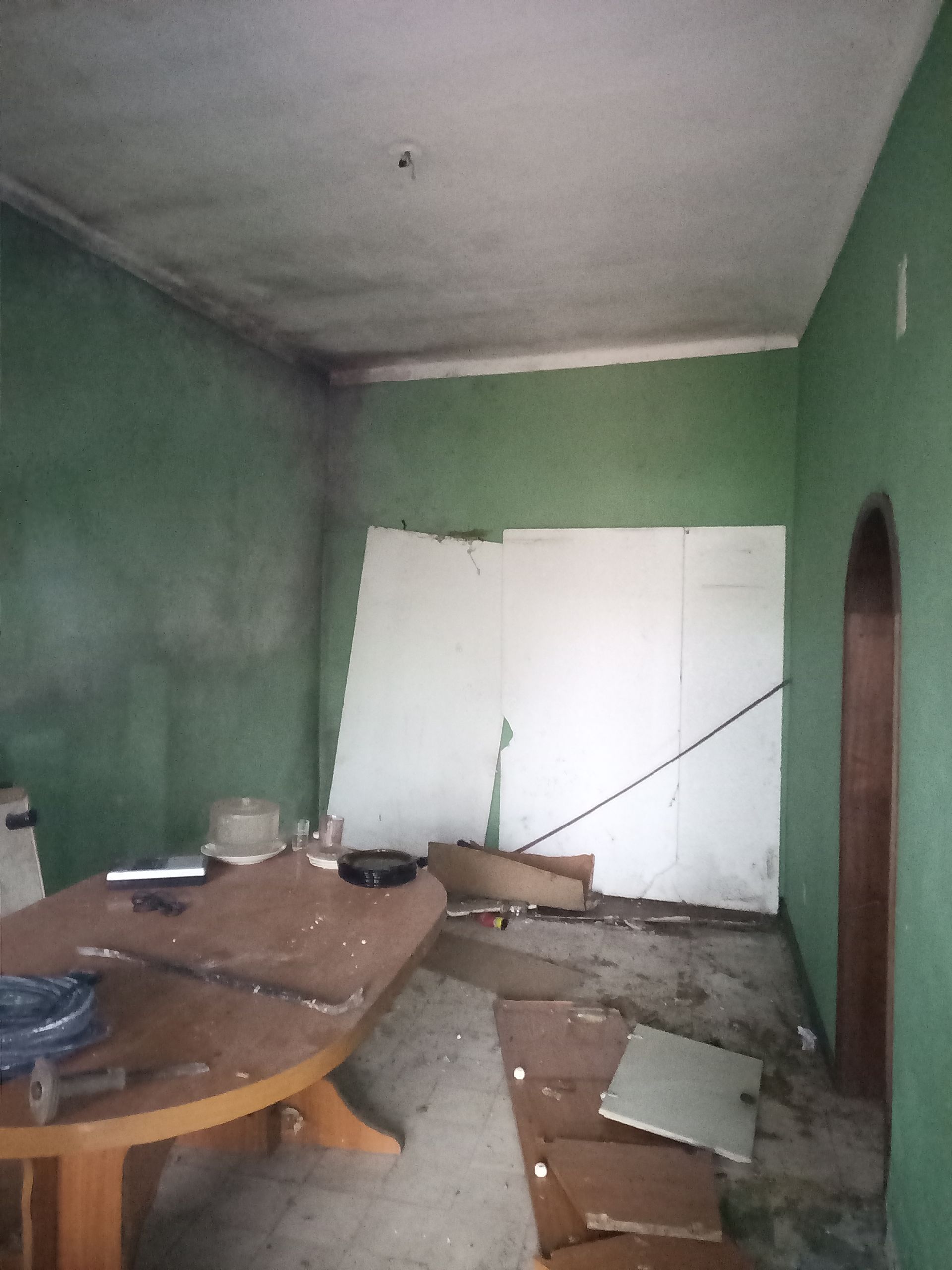 Quarto desarrumado com paredes verdes, painéis brancos danificados e detritos no chão, mesa oval.