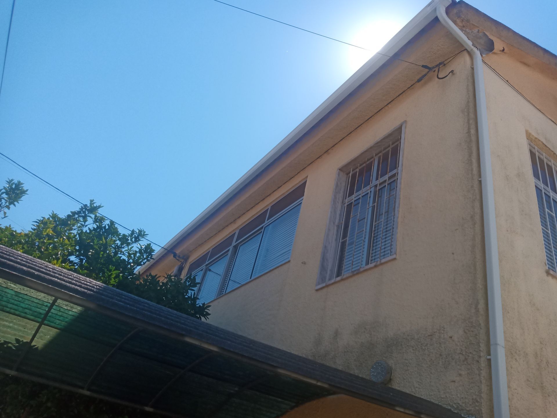 Prédio bege com calhas e janelas brancas contra um céu azul, com sol brilhando.