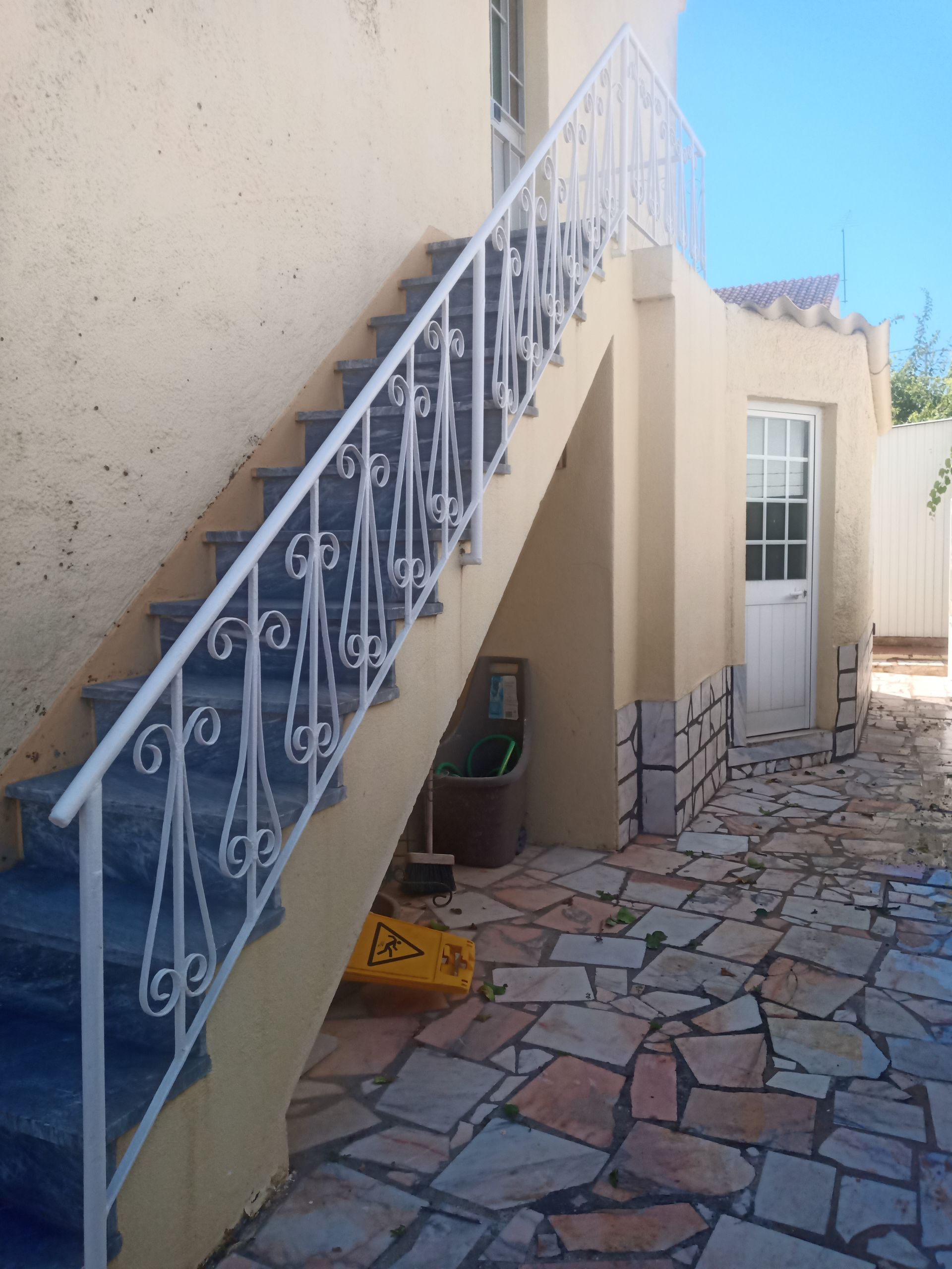 Escada externa com corrimão branco, que leva a uma porta. Paredes bege, pátio de pedra.