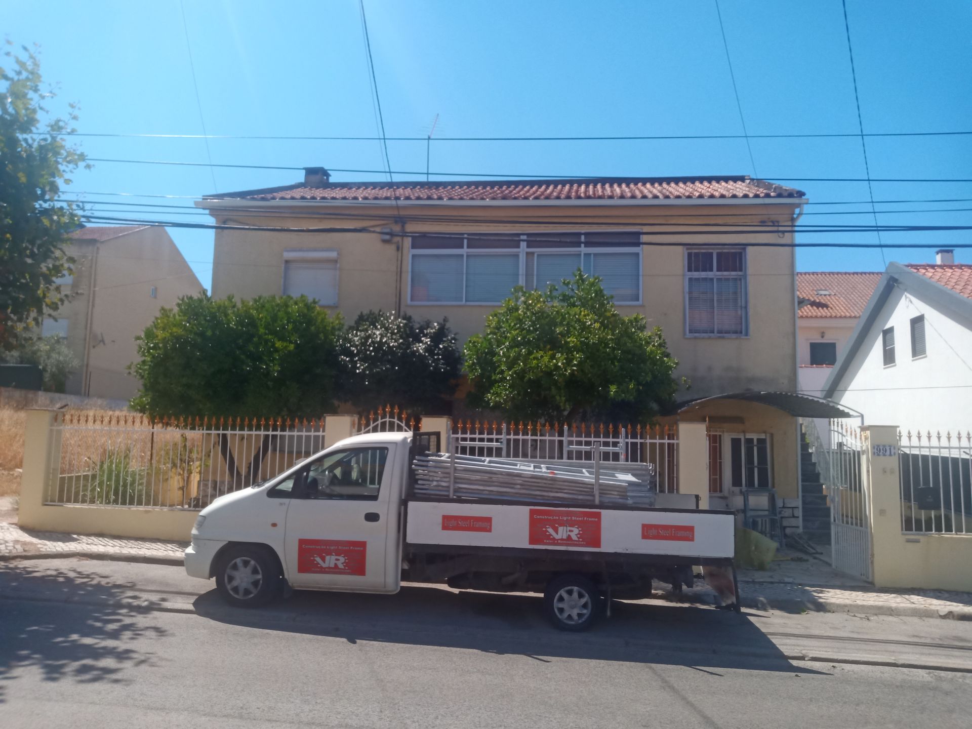 Caminhão branco estacionado em frente a um prédio de dois andares com cerca de madeira e árvores em um dia ensolarado.