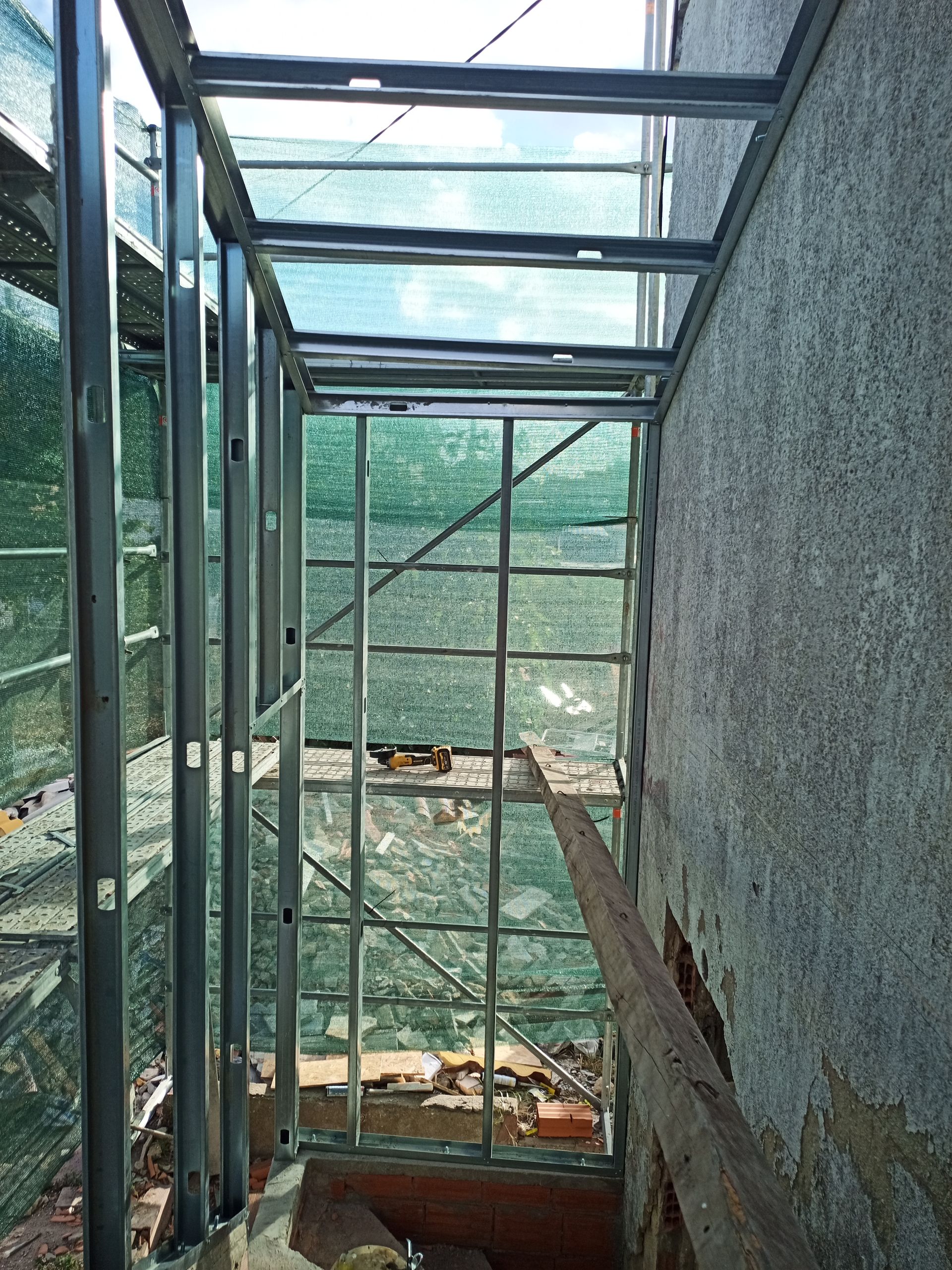 Estrutura metálica com painéis de vidro em construção ao lado de uma parede de concreto bruto.